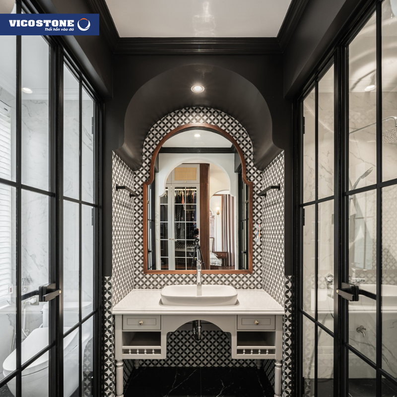 Khu vực lavabo với mặt bàn ốp đá VICOSTONE Misterio BQ8815 trắng tinh khôi, tạo thêm điểm nhấn bởi đường viền gỗ cong của gương và tường ốp gạch bông họa tiết hình học, thể hiện sự giao thoa giữa Đông - Tây đặc trưng
