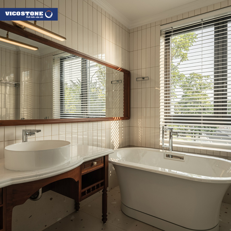 Phòng tắm tràn ngập ánh sáng tự nhiên với bồn tắm độc lập và mặt bàn lavabo ốp đá VICOSTONE Misterio BQ8815, kết hợp gương chữ nhật khung gỗ mang đến không gian thư thái, gần gũi thiên nhiên theo tinh thần Indochine (Nguồn: Biệt thự song lập Ecopark Ecorivers Hải Dương do kiến trúc sư Ngô Nguyễn Trung từ 3003 Architecture & Interior Design thiết kế).